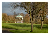 A park setting with some trees, grass and a footpath in the foreground. Towards the background is a bandstand with a green copper roof that is a dome shape, rising to a point at the centre. There are more trees behind the bandstand.