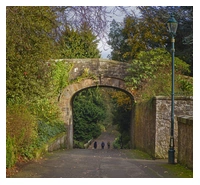 A stone bridge over a footpath. The bridge has an archway and there is a footpath running from the forefound, going through the arch and sloping down towards the background of the image. There are three small figures walking along the footpath. The arch is surrounded by trees and greenery, with a decorative lampost sited before it.