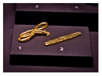 Two golden objects. One is a cigar shaped ingot. The other is a thin strip of gold twisted over on itself a couple of times with the ends hooked over each other.