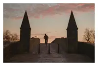 The pic shows a cobblestone path which forms part of an old bridge. There are two pointed pillars, along with some bollards, as the bridge arches over the (unseen) river. A cyclist is visible beyond the bollards. The sky has red and pink clouds.