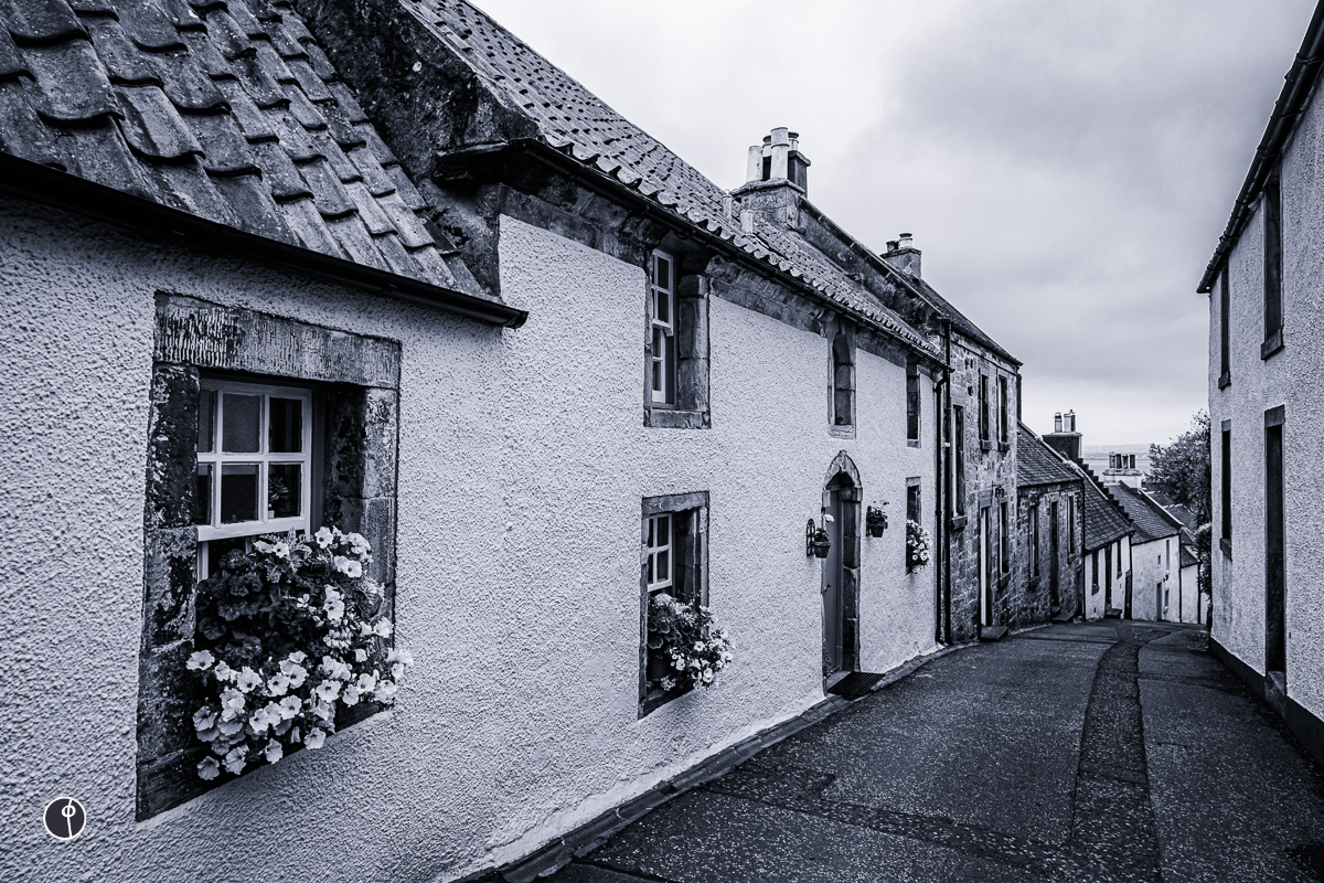 A row of old cottages lining on both sides of a narrow road. The closest cottage has a flower basket at the window.